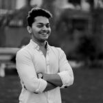 Black and white of young cheerful guy in classic shirt standing with crossed arms in park and smiling